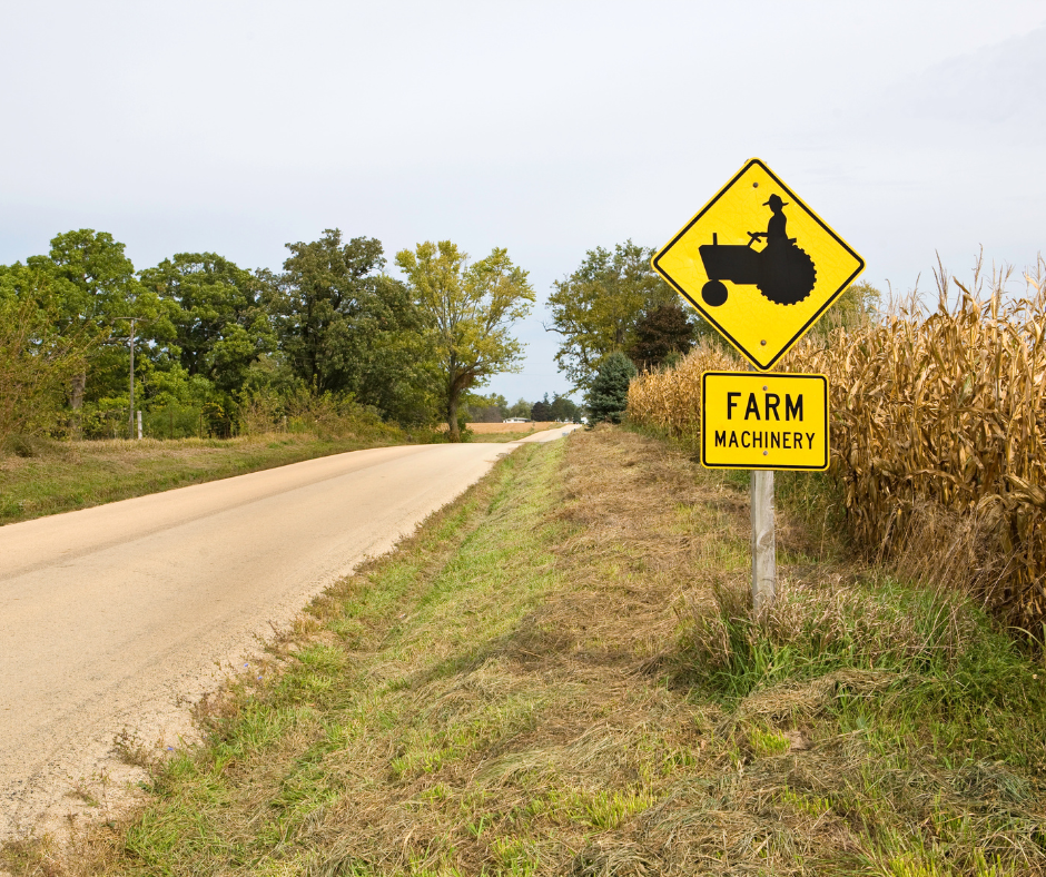 A photo of a rural roadway on a sunny spring day. The road is unmarked near a grassy ditch with a yellow farm machinery crossing road sign.